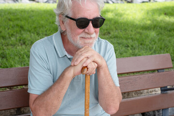 Smiling white haired older 70s man with walking disability sitting in a bench in the park holding...