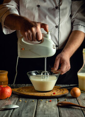 A person uses a hand mixer to blend cream in a glass bowl on a wooden table. Nearby ingredients...