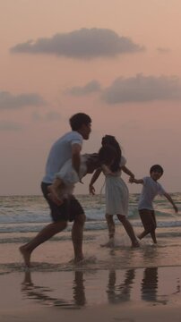 Vertical Screen : Asian young happy family enjoy vacation on beach in evening. Dad, mom and kid relax play together near sea silhouette sunset. Lifestyle travel holiday vacation summer. Slow motion.
