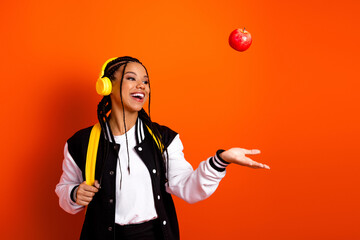 Happy teenage girl tossing an apple with headphones and a stylish jacket against a vibrant orange background