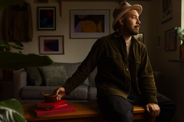 Portrait of a caucasian man with a beard wearing a green jacket sitting in a cosy living room holding a cup of coffee.
