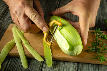 Hands hold a peeler, skillfully removing the skin from a green vegetable marrow on a wooden board....