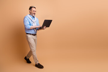 Elegant mature businessman walks confidently holding laptop against beige background exuding professional style
