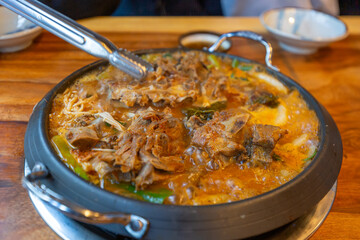 Korean food gamjatang, A bowl of food with meat and vegetables in a black pot. The meat is cut into pieces and the vegetables are chopped. The dish is served on a wooden table