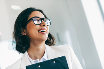 Professional businesswoman in formal attire smiling indoors with clipboard during work, expressing...