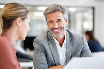 A professional man in a gray blazer smiles warmly while conversing with a woman in an office setting.