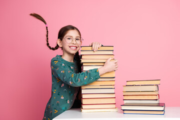 Smiling girl with braided hair holding a stack of book while posing against a bright pink background