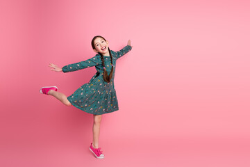 Cute joyful young schoolgirl posing against a pink background in a colorful dress and showing...