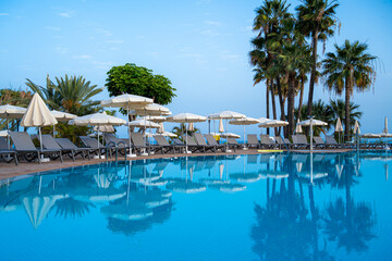 A pool with many umbrellas and lounge chairs. The pool is surrounded by palm trees. Scene is...