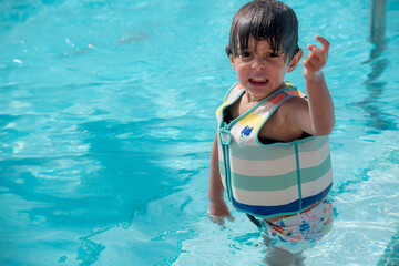 A young child is in a pool wearing a life jacket. The child is crying and pointing at something in the water