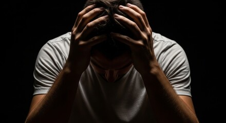 Stressed man holding head in hands against dark background, expressing mental fatigue, depression or overwhelming emotions in dramatic low-key lighting