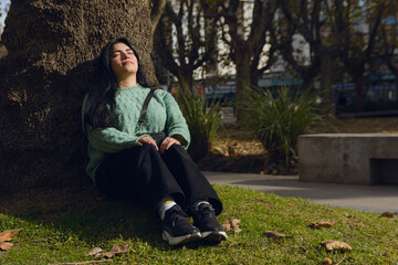 Young Venezuelan woman sitting in the park leaning against a tree under the sun