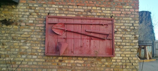 Vintage fire fighting tools displayed on a red wooden board, mounted on a brick wall. Axe, shovel, bucket, and hook. Fire shield. Displayed outdoors against a brick wall. Red wooden fire shield.