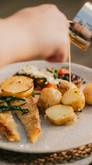A plate of food with a person pouring sauce on it. The plate has a variety of food items including potatoes, fish, and vegetables. The sauce is being poured over the fish and vegetables