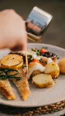 A person is pouring sauce over a plate of food. The plate contains fish, potatoes, and vegetables