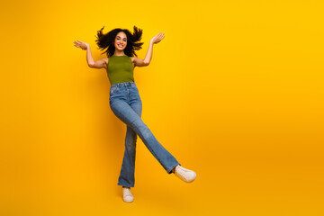 Young woman with curly hair dancing joyfully in trendy outfit on vibrant yellow background during a...