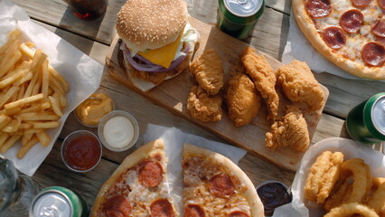 Top view of a wooden table filled with junk food, including cheeseburgers, pepperoni pizza slices, crispy fried chicken, french fries, onion rings, and dipping sauces, soft drink cans.