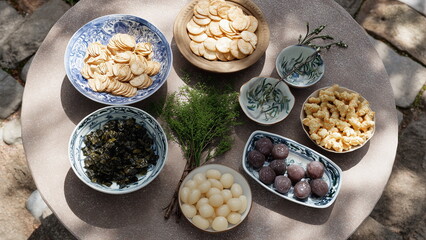 Top view of a round table with Asian-style tidbits including rice crackers, dried cuttlefish, wasabi peas, seaweed snacks, and sweet preserved plums, traditional porcelain dishes.