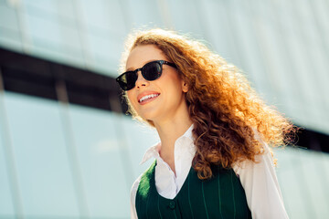 Confident businesswoman with curly red hair walking outdoors in a modern urban setting while...