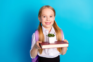 Cheerful schoolgirl with blonde hair holding a book and plant on blue background representing...