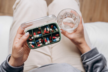 woman holding a glass of water in one hand and a variety of colorful capsules in the other, preparing to take medication, showcasing health and wellness routine in a cozy indoor setting