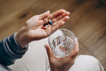 woman holding a glass of water in one hand and a variety of colorful capsules in the other, preparing to take medication, showcasing health and wellness routine in a cozy indoor setting