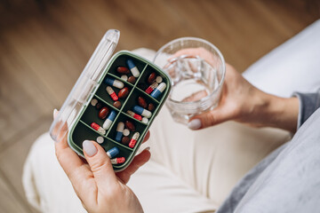woman holding a glass of water in one hand and a variety of colorful capsules in the other, preparing to take medication, showcasing health and wellness routine in a cozy indoor setting