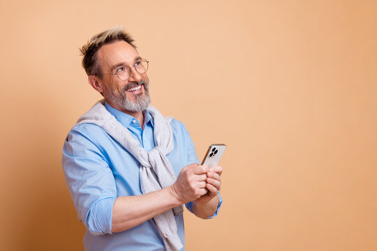 Elegant mature businessman in casual attire poses with smartphone against beige backdrop, showcasing stylish lifestyle and corporate charm
