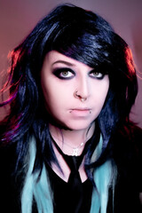 Fototapeta premium Alternative girl with dark makeup, striped sleeves, and facial piercings posing indoors under moody red and purple lighting.