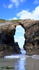 A stunning natural arch carved into towering sea cliffs on a sandy beach, with the ocean visible through the opening. 