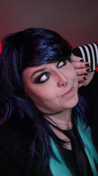 Alternative girl with dark makeup, striped sleeves, and facial piercings posing indoors under moody red and purple lighting.