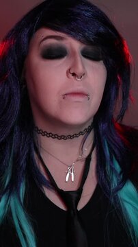 Alternative girl with dark makeup, striped sleeves, and facial piercings posing indoors under moody red and purple lighting.