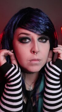 Alternative girl with dark makeup, striped sleeves, and facial piercings posing indoors under moody red and purple lighting.