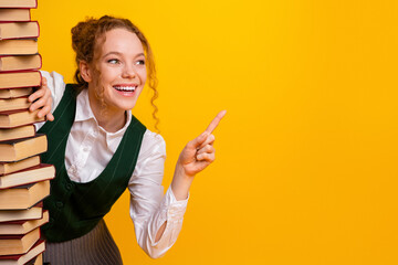 Cheerful young woman leaning on a stack of books, pointing and smiling against a bright yellow...