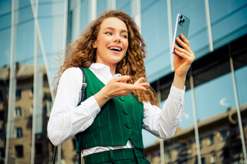 Confident young businesswoman taking a cheerful selfie outdoors in a modern urban setting, wearing...