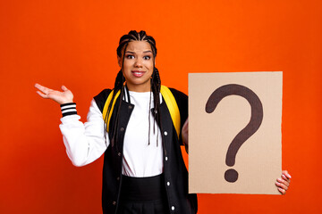 Young woman with a playful expression holding a sign with a question mark against a vibrant orange...