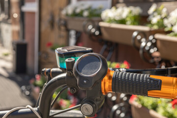 Fototapeta premium Detailed image of a black bicycle handlebar with an orange grip, bell, and blurred background of bright flowers.