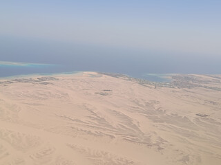 High-altitude photograph showing a vast desert landscape meeting the clear blue waters of the sea. Taken from an airplane window, the image captures sandy textures