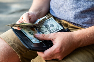 Counting Cash – Man with Black Wallet and US Dollar Banknotes. A close-up view of a man counting U.S. dollar banknotes from a black leather wallet while seated on a bench