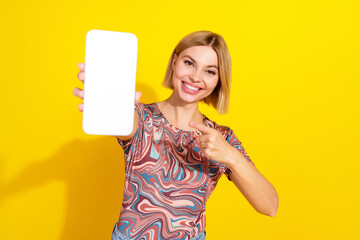 Young trendy woman holding a blank smartphone against a vibrant yellow backdrop, pointing and...