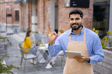 A smiling Hindu waiter wearing an apron at an outdoor restaurant, holding a tablet and giving an okay gesture.