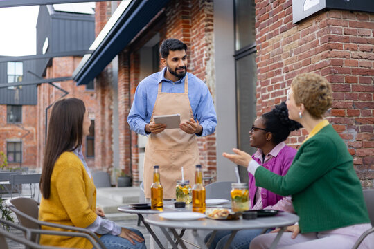 A waiter takes an order on a tablet at an outdoor cafe. Three women sit at the table, socializing.