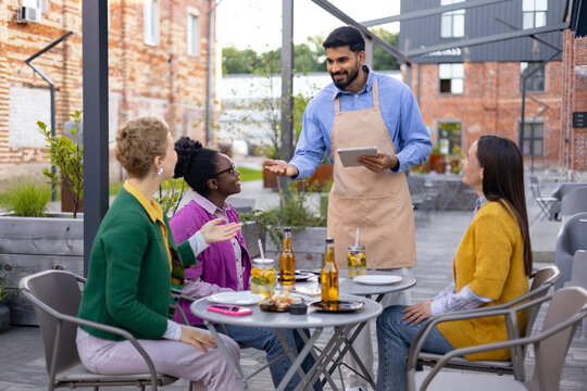 A waiter takes an order from three women seated at an outdoor restaurant table. They are enjoying drinks and conversation.