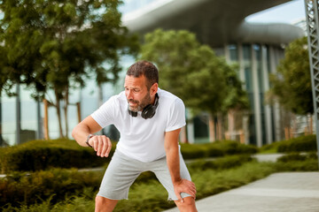 Focused runner checks watch during brisk morning jog in urban park surrounded by modern architecture and greenery
