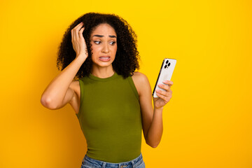 Young stylish woman in green top holding smartphone with confused expression against bright yellow...