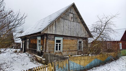 An old wooden house with a slate roof stands on a concrete foundation. There is a wooden fence, trees and houses around. In winter, everything is covered with snow. Cloudy weather and gray sky