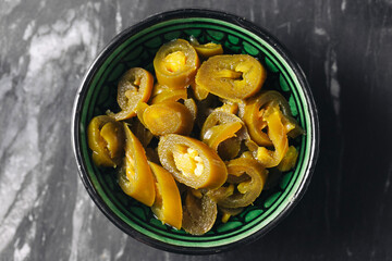 Pickled jalapeno chili slices in ceramic bowl. Spicy green peppers on dark marble background. Tangy preserved vegetable condiment for cooking or snacking. Mexican flavor and texture close-up