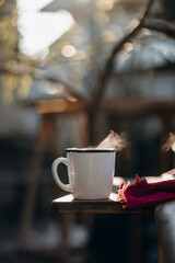 A white cup of hot coffee emits steam in warm morning sunlight. Set on a wooden table in a peaceful garden, alongside a clear glass coffee brewer, capturing a serene cottagecore vibe.