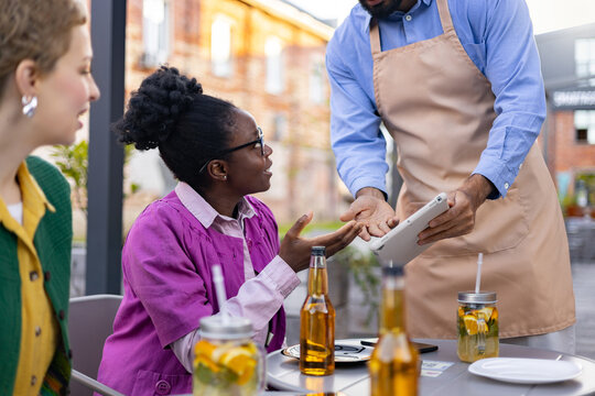 A diverse group of people enjoying a meal outdoors. A waiter takes an order on a tablet. - Powered by Adobe
