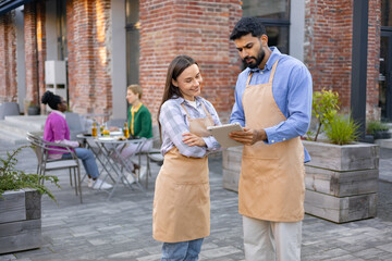 A diverse pair of restaurant staff using a tablet while discussing something outside a brick...